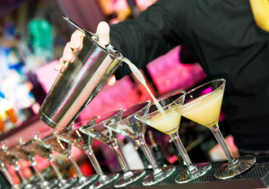 Barman hand with shake mixer pouring cocktail into glasses arranged in a line